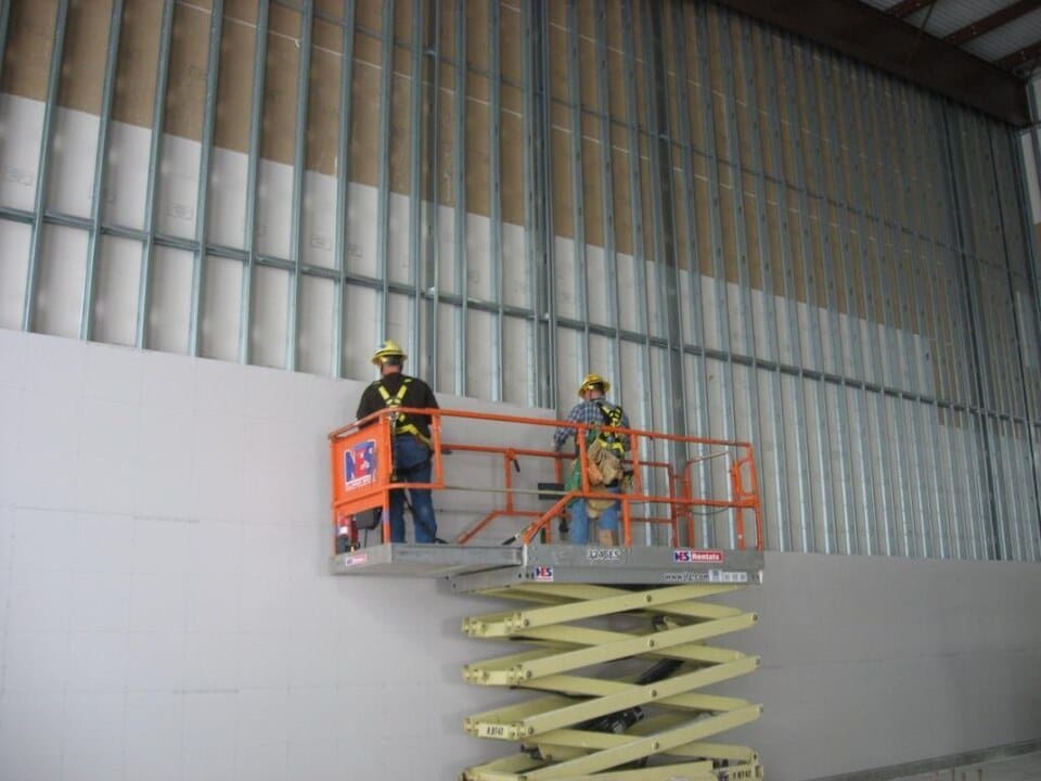 Workers installing wallboard from a scissor lift inside a building