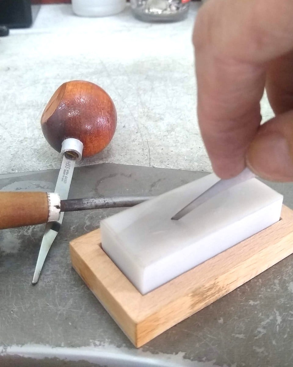 A hand sharpens an engraving tool on an Arkansas stone at a working bench.