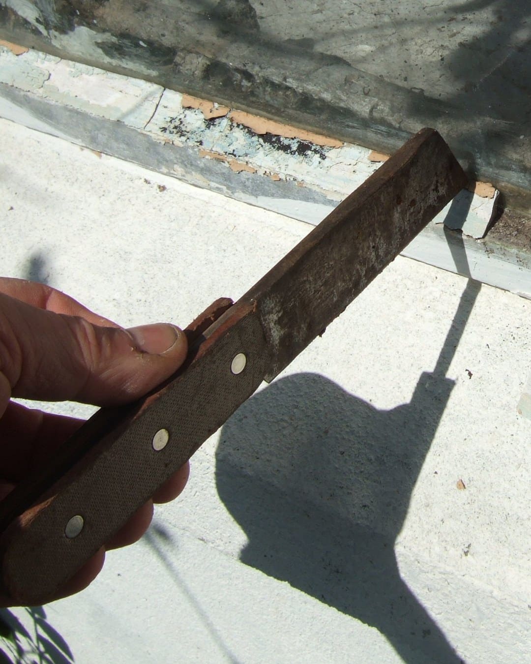 A hand using a hacking knife to remove old glazing putty from a window.